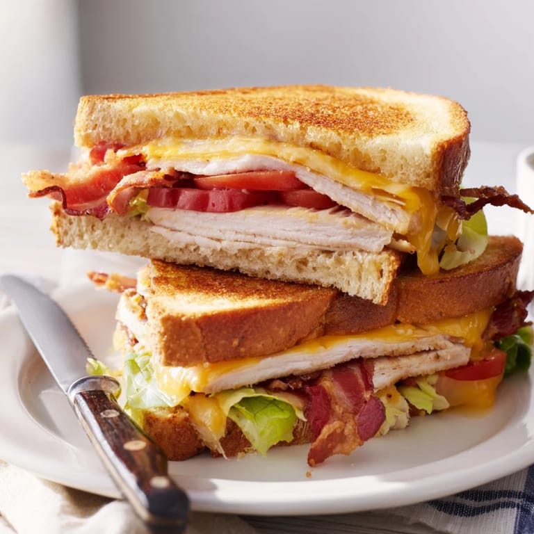 Ein heißer Turkey Club Grilled Cheese aus einer Butterpfanne, belegt mit Schinkenspeck, Truthahnscheiben, Romana-Salat und Tomaten in honigfarbenem Sauerteigbrot.